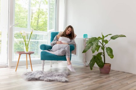 Photo for Beautiful young pregnant woman relaxing at home and expecting her baby; pregnant woman sitting in an armchair, holding her belly and smiling - Royalty Free Image