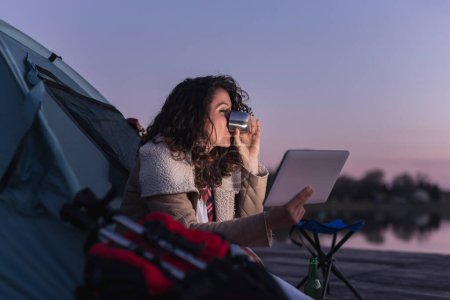 Foto de Mujer joven acampando junto al lago, sentada en la entrada de la tienda, bebiendo café caliente y leyendo las noticias usando tableta - Imagen libre de derechos