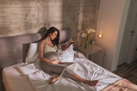 Foto de Hermosa mujer embarazada vistiendo camisón, acostada en la cama y relajándose en casa por la mañana, leyendo un libro - Imagen libre de derechos
