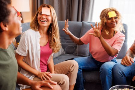 Foto de Grupo de alegres jóvenes amigos divirtiéndose en casa reuniéndose jugando palabras adivinando juego charadas y pantomima - Imagen libre de derechos