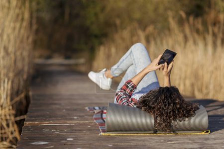 Téléchargez les photos : Femme allongée sur les quais du lac, se détendre après une longue journée de randonnée dans la nature, en utilisant un téléphone intelligent - en image libre de droit