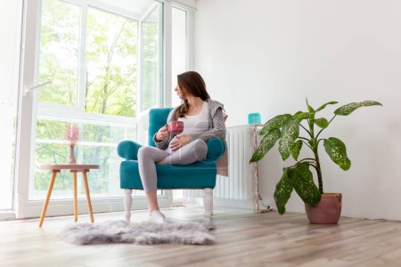Foto de Mujer embarazada relajándose en casa, sentada en un sillón junto a la ventana, bebiendo una taza de té caliente y abrazándose el vientre, deseando conocer a su bebé - Imagen libre de derechos