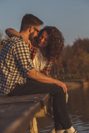 Foto de Hermosa pareja joven enamorada sentada en los muelles de un lago, abrazándose y disfrutando de una hermosa puesta de sol sobre el lago - Imagen libre de derechos