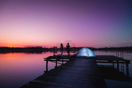 Téléchargez les photos : Couple campant sur les quais du lac au crépuscule, debout à côté d'une tente profitant de belles nuits d'été et les couleurs du ciel juste après le coucher du soleil - en image libre de droit