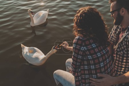 Foto de Vista de ángulo alto de pareja joven enamorada sentada en los muelles del lago, abrazando, alimentando cisnes y disfrutando de un hermoso día soleado en la naturaleza - Imagen libre de derechos