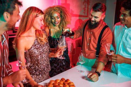 Téléchargez les photos : Groupe d'amis joyeux s'amuser à une fête, l'ouverture d'une bouteille de vin tout en célébrant les amis anniversaire ensemble à la maison - en image libre de droit