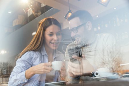 Téléchargez les photos : Portrait d'un jeune couple amoureux assis à une table de restaurant, s'amusant lors d'un rendez-vous, utilisant un téléphone intelligent et buvant du café - en image libre de droit