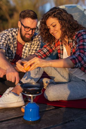 Téléchargez les photos : Couple camper au bord du lac, cuisiner des œufs pour le dîner et s'amuser dans une aventure nature en plein air - en image libre de droit