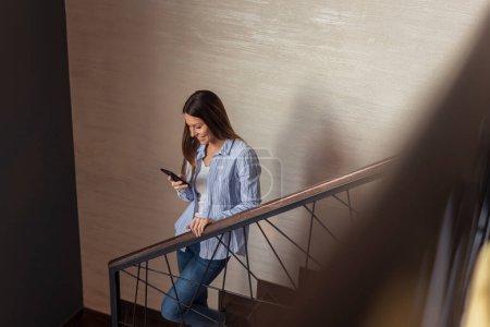 Foto de Hermosa mujer joven de pie en una escalera, sonriendo mientras escribe un mensaje de texto en un teléfono inteligente - Imagen libre de derechos