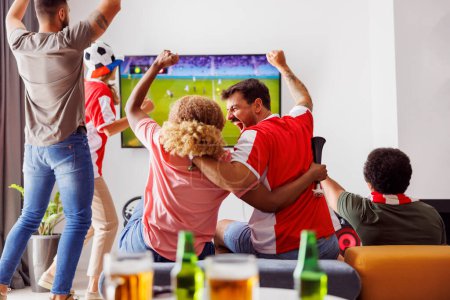 Foto de Grupo de jóvenes amigos divirtiéndose viendo partidos de fútbol en la televisión, bebiendo cerveza y animando; fanáticos del fútbol viendo partidos en casa celebrando después de que su equipo anotara un gol - Imagen libre de derechos