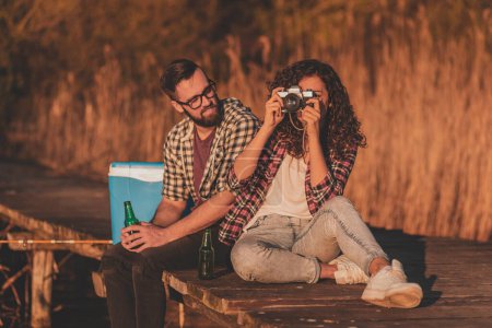 Foto de Hermosa pareja joven enamorada sentada en los muelles del lago, bebiendo cerveza y tomando fotos, disfrutando de un hermoso día de otoño en la naturaleza - Imagen libre de derechos