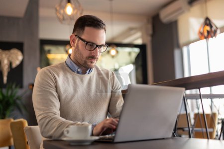 Téléchargez les photos : Homme d'affaires assis à une table de restaurant, buvant du café et travaillant sur un ordinateur portable - en image libre de droit