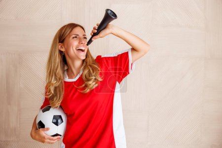 Téléchargez les photos : Portrait d'une fan de football féminine portant un maillot de sport, tenant un ballon de football et soufflant du cor de football tout en soutenant son équipe nationale au championnat du monde - en image libre de droit