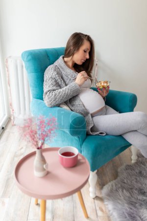 Téléchargez les photos : Belle femme enceinte assise dans un fauteuil, mangeant une salade mixte de légumes de saison ; concept de grossesse et d'alimentation saine - en image libre de droit