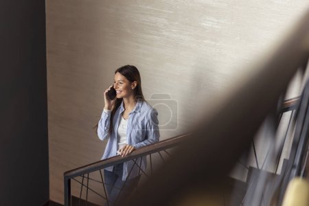 Foto de Hermosa mujer joven de pie en una escalera, sonriendo mientras tiene una conversación telefónica - Imagen libre de derechos