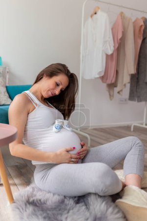 Photo for Beautiful young pregnant woman relaxing at home, expecting her baby; pregnant woman sitting on the floor next to an armchair, holding tiny baby shoes on her belly and smiling - Royalty Free Image