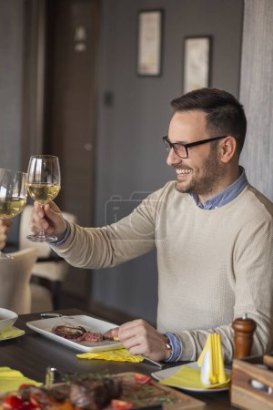 Téléchargez les photos : Jeune couple amoureux à un rendez-vous, assis à une table de restaurant, lever des verres de vin et porter un toast tout en déjeunant - en image libre de droit
