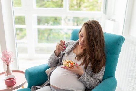 Foto de Hermosa mujer embarazada sentada en un sillón, comiendo ensalada mixta de verduras de temporada; concepto de embarazo y dieta saludable - Imagen libre de derechos