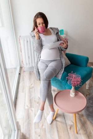 Téléchargez les photos : Belle femme enceinte relaxante à la maison, assise dans un fauteuil, buvant du thé et surfant sur le net sur une tablette - en image libre de droit