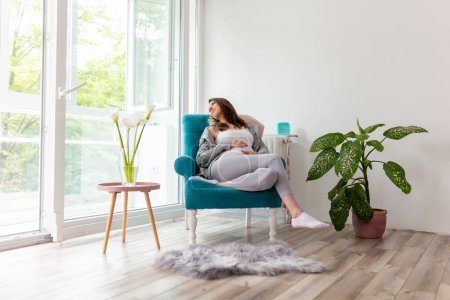 Foto de Hermosa joven embarazada relajándose en casa y esperando a su bebé; mujer embarazada sentada en un sillón, sosteniendo su vientre, mirando por la ventana y sonriendo - Imagen libre de derechos