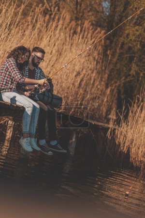 Foto de Pareja enamorada sentada en los muelles del lago, pescando y disfrutando de un hermoso día soleado de otoño en la naturaleza - Imagen libre de derechos