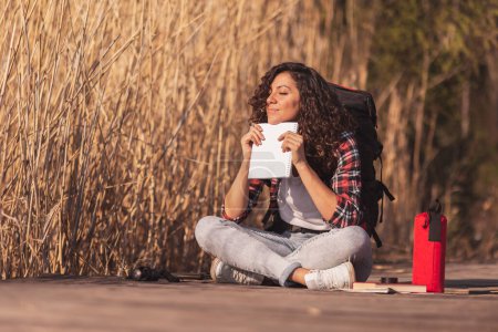 Photo for Beautiful young woman sitting on the wooden lake docks, enjoying a sunny autumn day in nature, holding a notebook, pensive and relaxed - Royalty Free Image