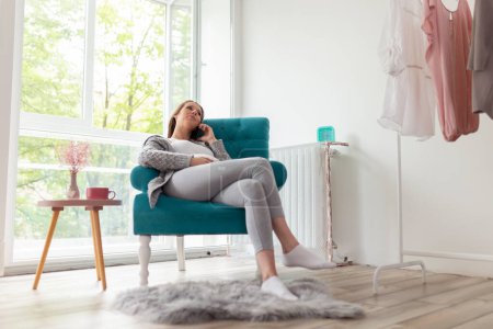 Foto de Hermosa mujer embarazada sentada en un sillón, relajándose en casa, tomando té y teniendo una conversación telefónica - Imagen libre de derechos