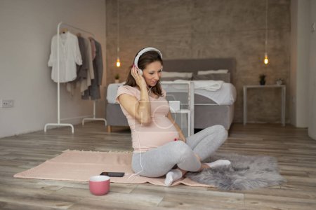 Téléchargez les photos : Belle femme enceinte profitant de son temps libre à la maison, écoutant la musique, buvant du thé et se relaxant - en image libre de droit