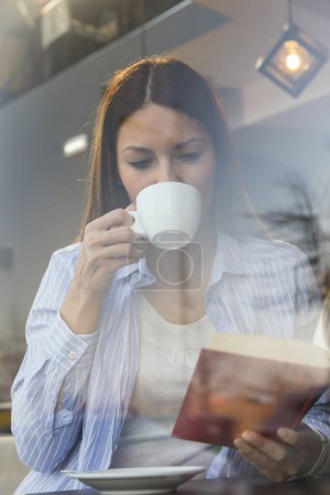 Schöne junge Frau sitzt an einem Restauranttisch, trinkt Kaffee und liest ein Buch