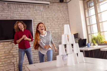 Foto de Gente de negocios divirtiéndose en la oficina, jugando juegos de oficina mientras están en un descanso, derribando una pirámide de copas de plástico lanzando una bola de papel - Imagen libre de derechos