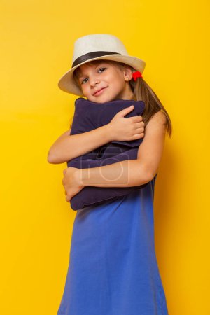 Photo for Beautiful little girl wearing blue dress and summer hat, hugging a travel pillow, getting ready for summer vacations , isolated on yellow colored background - Royalty Free Image