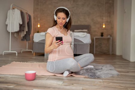 Foto de Hermosa mujer embarazada disfrutando de su tiempo libre en casa, eligiendo una canción de una lista de reproducción de teléfonos inteligentes, escuchando la música y relajándose - Imagen libre de derechos