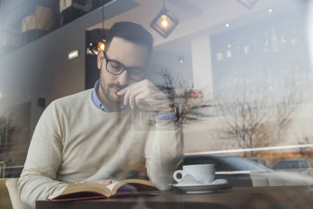 Foto de Retrato de un hombre apuesto y pensativo sentado en la mesa de un restaurante, tomando café y leyendo un libro - Imagen libre de derechos