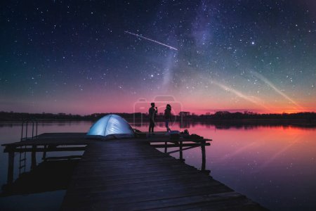 Foto de Combinación de alta ISO, imagen ruidosa de un cielo estrellado e imagen de una pareja acampando en los muelles del lago al atardecer, de pie junto a una tienda de campaña disfrutando de una hermosa noche de verano - Imagen libre de derechos
