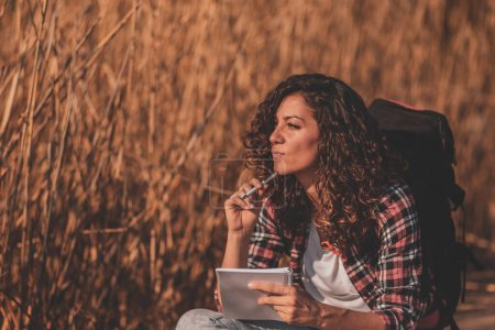 Téléchargez les photos : Jeune écrivaine portant un sac à dos assis sur les quais d'un lac en bois par une journée ensoleillée d'automne, prenant des notes dans un planificateur, faisant un concept pour un nouveau livre - en image libre de droit