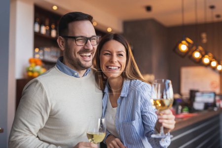 Photo for Young couple in love on a date, standing next to a restaurant counter, raising glasses of wine and making a toast while waiting for a table - Royalty Free Image