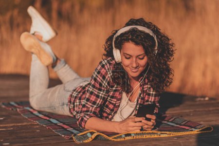 Téléchargez les photos : Femme portant un casque couché sur les quais du lac, utilisant un téléphone intelligent, choisissant une chanson dans une playlist, écoutant la musique un jour d'automne ensoleillé - en image libre de droit