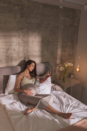 Téléchargez les photos : Belle femme enceinte en chemise de nuit, couchée au lit et relaxante à la maison le matin, lisant un livre - en image libre de droit