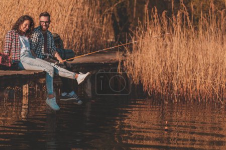 Foto de Pareja enamorada sentada en los muelles del lago, pescando y disfrutando de un hermoso día soleado de otoño en la naturaleza - Imagen libre de derechos