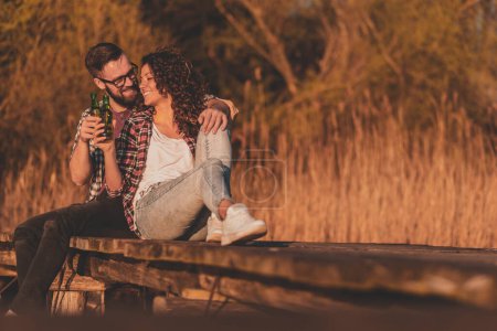 Foto de Pareja sentada en los muelles del lago, disfrutando de la hermosa puesta de sol de otoño, bebiendo cerveza y divirtiéndose - Imagen libre de derechos