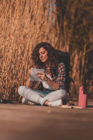 Téléchargez les photos : Jeune biologiste portant un sac à dos assis sur les quais d'un lac en bois par une journée ensoleillée d'automne, écrivant dans un carnet pendant une sortie de travail sur le terrain - en image libre de droit