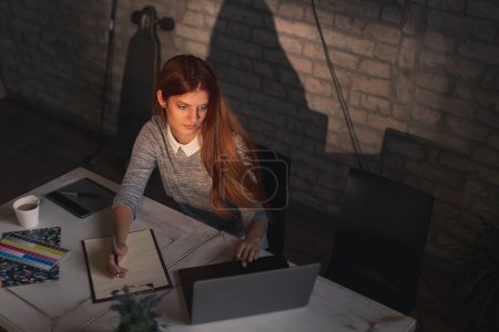Photo for High angle view of woman telecommuting, working on a laptop computer and taking notes in a planner - Royalty Free Image