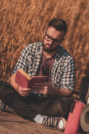 Téléchargez les photos : Homme assis sur les quais du lac en bois, profitant d'une belle journée d'automne dans la nature et lisant un livre - en image libre de droit