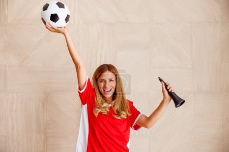 Téléchargez les photos : Fan de football féminin portant un maillot de sport, tenant un ballon de football et soufflant du cor de football tout en soutenant son équipe nationale lors d'une compétition internationale - en image libre de droit