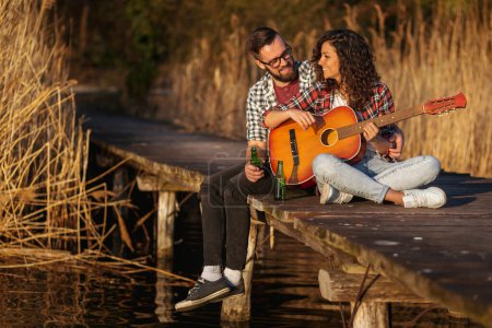 Téléchargez les photos : Couple assis sur les quais du lac, jouant de la guitare, chantant et buvant de la bière, profitant d'une journée dans la nature et se relaxant - en image libre de droit