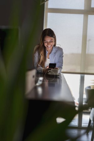 Téléchargez les photos : Portrait d'une belle jeune femme assise à une table de restaurant, buvant du café et tapant un message texte sur un smartphone - en image libre de droit