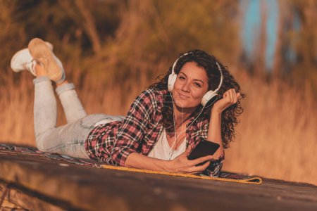 Téléchargez les photos : Femme portant un casque couché sur les quais du lac, utilisant un téléphone intelligent, choisissant une chanson dans une playlist, écoutant la musique un jour d'automne ensoleillé - en image libre de droit