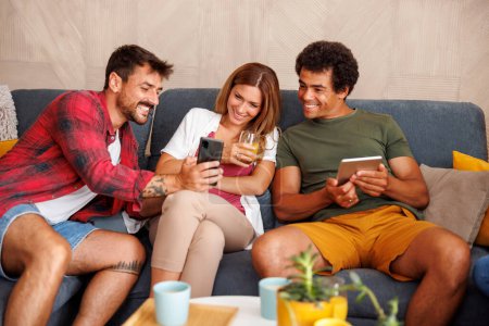 Téléchargez les photos : Groupe d'amis utilisant un téléphone intelligent et une tablette, surfant sur le net et s'amusant à passer du temps libre ensemble à la maison - en image libre de droit