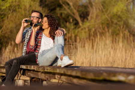 Téléchargez les photos : Couple assis sur les quais du lac, profiter d'un magnifique coucher de soleil d'automne, boire de la bière et s'amuser - en image libre de droit