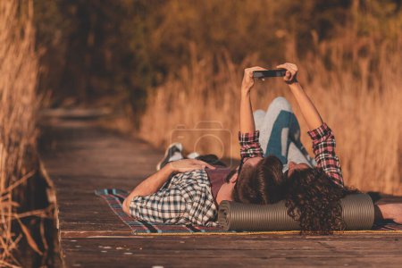 Téléchargez les photos : Beau couple amoureux couché sur une couverture de pique-nique aux quais du lac, prenant un selfie et s'amusant un jour d'automne ensoleillé - en image libre de droit